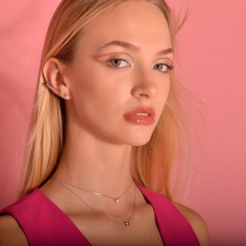 Modelo con cabello largo y rubio, luciendo joyería elegante, incluyendo pendientes y collares, frente a un fondo rosa. Su maquillaje resalta sus rasgos faciales, con labios brillantes y ojos bien definidos.