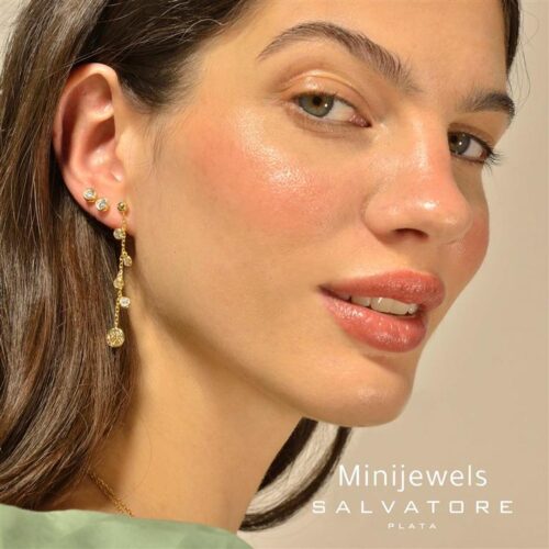 Mujer posando con aretes de la colección Minijewels de Salvatore, mostrando joyería elegante en un fondo neutral. Su maquillaje resalta su belleza natural y los detalles de los aretes.