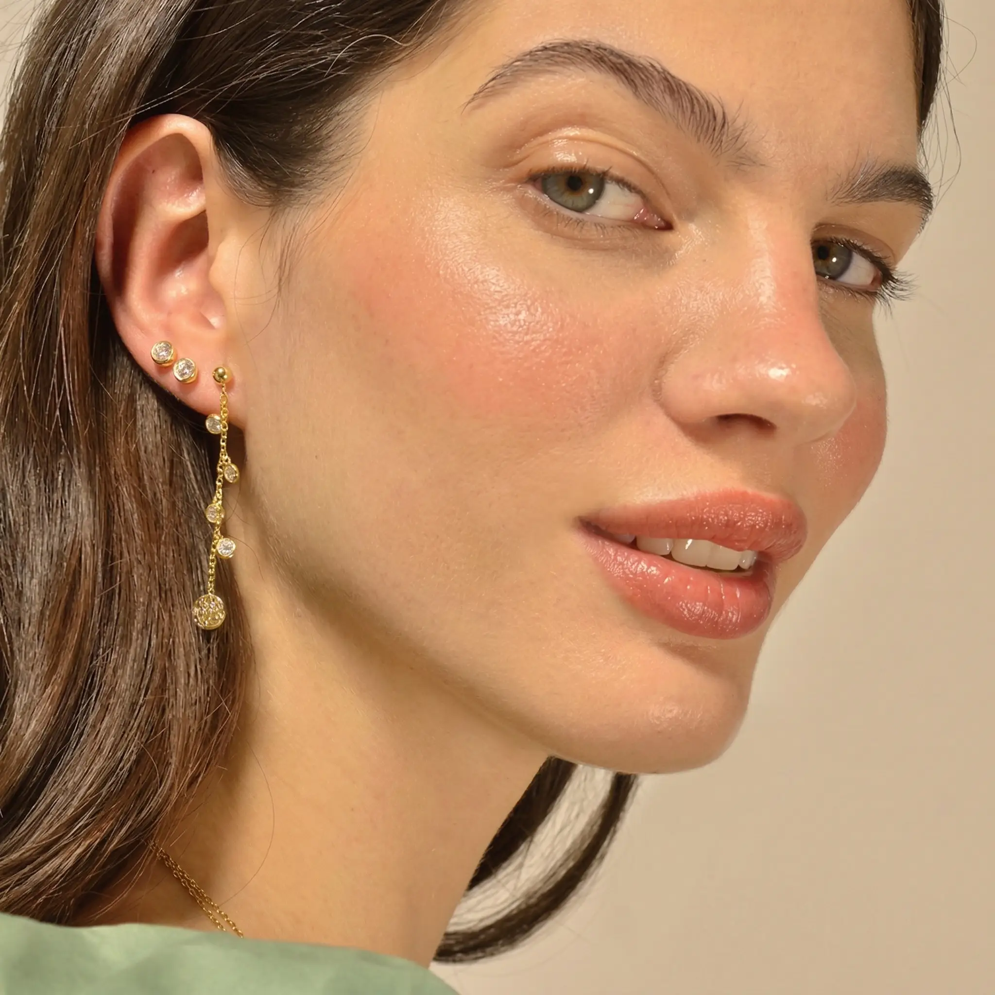 Mujer joven con pendientes de oro y piedras brillantes, mostrando un maquillaje natural y piel luminosa, con fondo neutro.