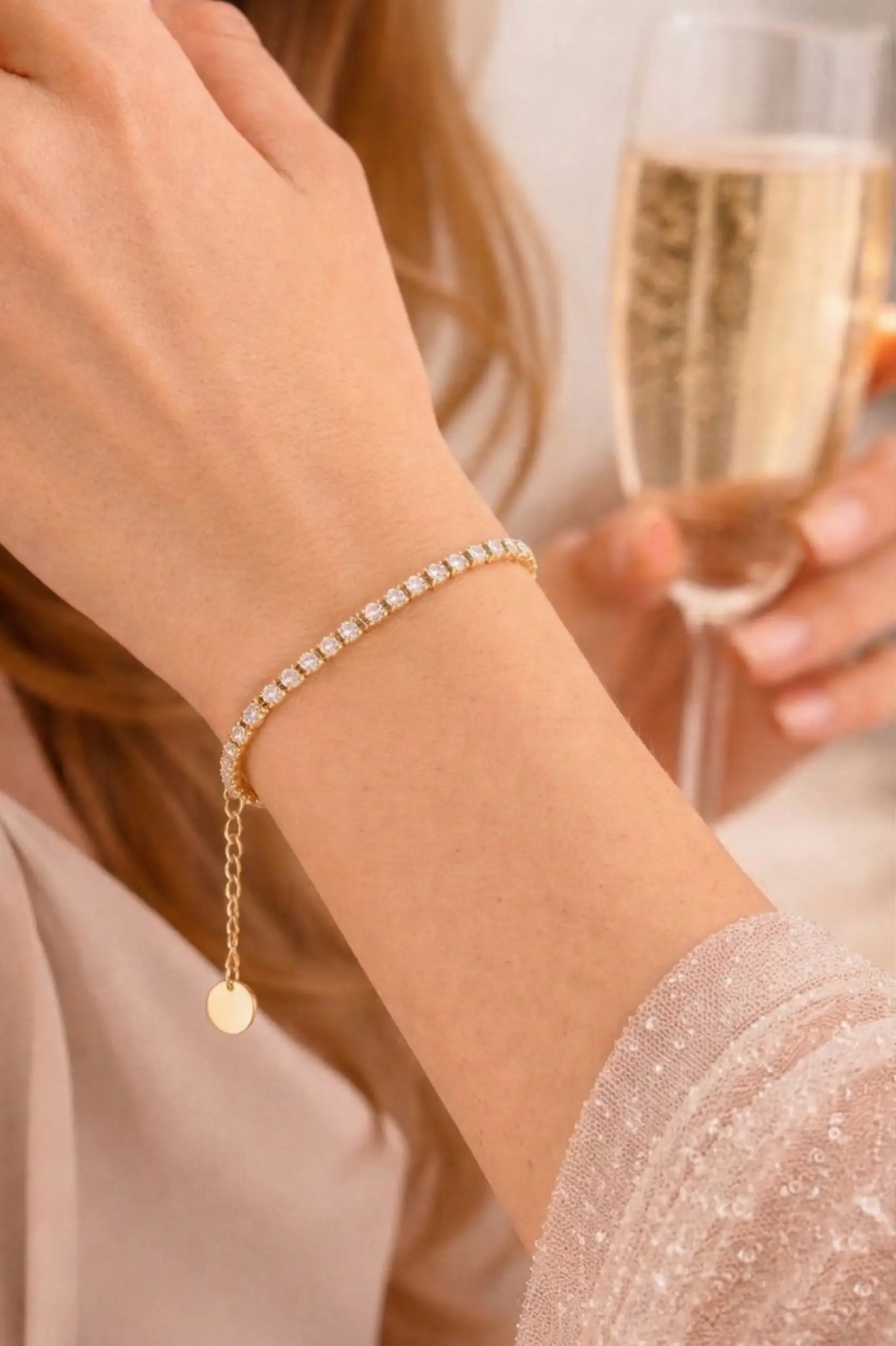 Pulsera de oro con piedras brillantes en una muñeca, mientras una mano sostiene una copa de champán. Ideal para ocasiones elegantes y celebraciones.