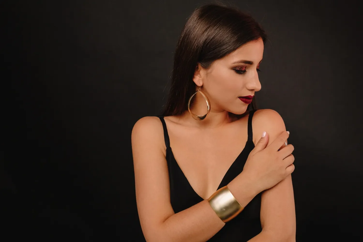 Mujer con cabello largo y lacio, usando un top negro, pendientes grandes y una pulsera dorada, posando con un fondo negro. Su maquillaje incluye sombras brillantes y labios oscuros, creando un look elegante y sofisticado.
