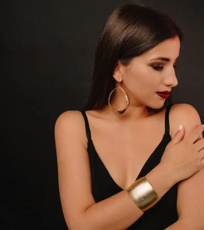 Mujer con cabello largo y lacio, usando un top negro, pendientes grandes y una pulsera dorada, posando con un fondo negro. Su maquillaje incluye sombras brillantes y labios oscuros, creando un look elegante y sofisticado.
