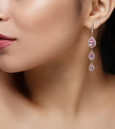 Pendiente largo de oro con piedras preciosas rosas, presentado en el rostro de una mujer con piel radiante y labios brillantes, fondo negro.